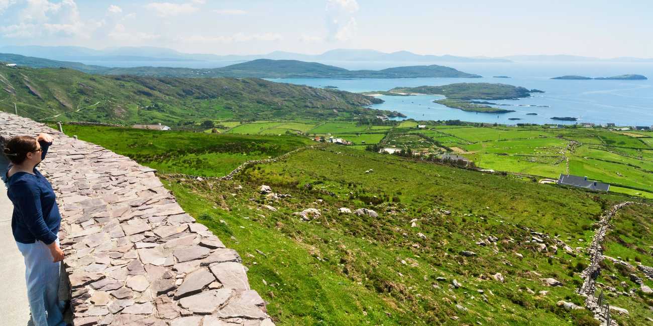 Explore the Kingdom of Kerry – The dramatic mountain and coastal scenery along the N70 highway in 2026 A traveler standing at a stone overlook admiring the panoramic green hills, traditional stone walls, and Atlantic ocean inlets at Coomakista Pass on the Ring of Kerry.